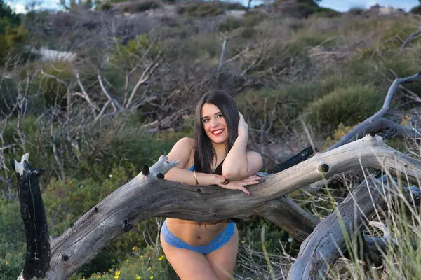 Genç bir kadın, güzel, esmer, mavi iç çamaşırlı, kameraya flört ederek bakıyor ve bir ağaç gövdesine yaslanmış gülümsüyor. Güzelliği kavra, doğayı, modayı, flörtü.