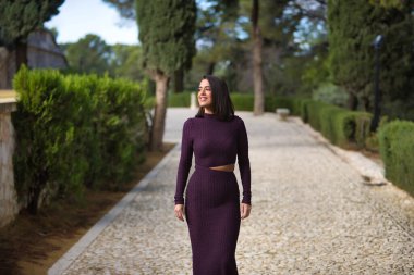 Genç, güzel, esmer, İspanyol bir kadın, mor bir elbise giyiyor, selvi ağaçlarıyla çevrili taş bir yolda tek başına yürüyor. Güzelliği, modayı, modayı kavra.