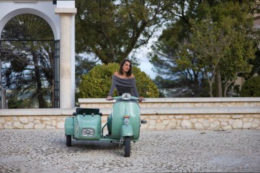 Genç, güzel, esmer, gri elbiseli İspanyol kadın sepetli antika bir motosiklet kullanıyor, dikkati dağınık görünüyor. Konsept motor, motosiklet, vintage, moda, güzellik.