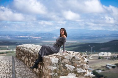 Genç, güzel, esmer, İspanyol kadın, gri bir elbise ve çizme giyiyor, gülümsüyor ve mutlu, taş bir duvarda oturuyor, arkasında bulutlardan ve kırsal alanlardan oluşan güzel bir manzara var..