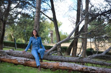 Genç, güzel, esmer, İspanyol bir kadın, kot elbise giyiyor, gülümsüyor ve mutlu, bir ağacın gövdesinde oturuyor, doğada. Kavram moda, güzellik, moda, kadınlık.
