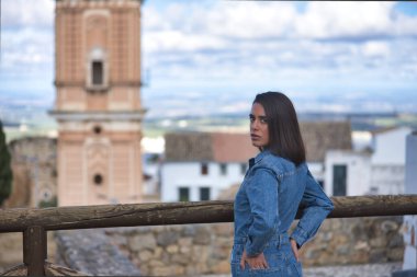 Genç, güzel, esmer, İspanyol kadın portresi, kot elbise giyiyor, utangaç ve içine kapanık, arkası dönük, arka ceplerinde, şehir manzarası olan.