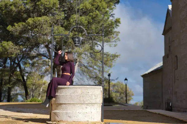 Genç, güzel, esmer, İspanyol bir kadın, mor bir elbise ve topuklu ayakkabı giyiyor, rahat ve sakin poz veriyor, eski bir su kuyusunda oturuyor. Güzelliği, modayı, modayı kavra.