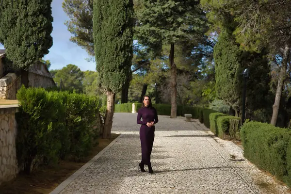 Genç, güzel, esmer, İspanyol bir kadın, mor bir elbise ve yüksek topuklu ayakkabı giyiyor, selvi ağaçlarıyla çevrili taş bir yolda tek başına yürüyor. Güzelliği, modayı, modayı kavra.