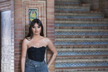 Genç, güzel, siyah dantelli ve kot pantolonlu esmer kadın, kameraya bakıyor, modern ve çağdaş, tuğla bir sütuna yaslanıyor..