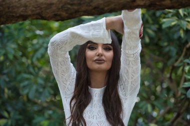 Genç, güzel, esmer bir kadının portresi, beyaz örgü bir elbise giymiş, kollarının oluşturduğu çerçeveden kameraya bakıyor, eli bir ağaç gövdesinde dinleniyor..