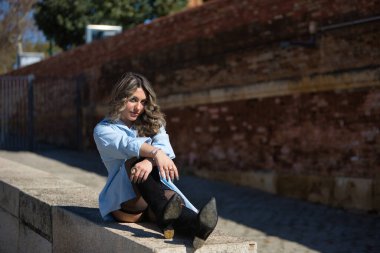 Genç, güzel, sarışın kadın, gömlek, deri etek ve botlar giyiyor, kameraya bakıyor, rahat ve sakin, taş bir duvarda oturuyor..