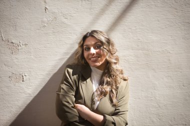 Genç, güzel, sarışın bir kadının portresi, yeşil bir ceket giymiş, kollarını kavuşturmuş, kameraya bakıyor, bağımsız ve güçlü, gülümseyen ve mutlu, duvara yaslanmış..