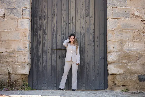 Genç, güzel, kahverengi saçlı bir kadın, açık gri bir ceket giyiyor, kameraya bakıyor, saçlarına dokunurken poz veriyor, kocaman, ahşap bir kapının ortasında..