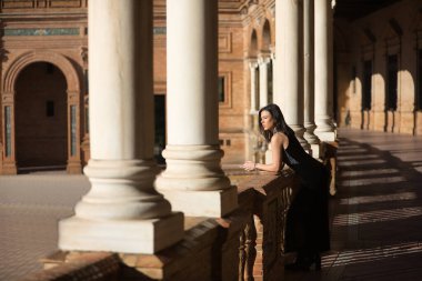 Genç, güzel, esmer, süslü siyah elbiseli bir kadın, tuğla duvara yaslanmış, Sevilla 'daki İspanya meydanına bakıyor. Güzellik, moda, moda, kadınlık, turizm kavramları.