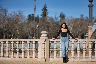 Genç, güzel, esmer bir kadın, siyah uzun kollu tişört ve kot pantolon giyiyor, saçları rüzgarda uçuşuyor, Seville 'de İspanya meydanında bir korkuluğa yaslanmış poz veriyor. Güzellik kavramı, kadınlık.