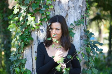 Siyah tişört ve kot pantolon giymiş, bir ağacın gövdesine yaslanmış ve doğayla bağlantılı, sarmaşığa dolanmış genç, güzel, koyu renk saçlı bir kadın. Güzellik kavramı, kadınlık, doğa, bitkiler, ağaçlar.