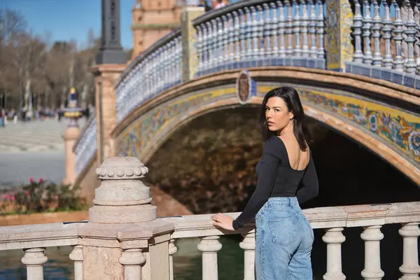 Genç, güzel, siyah uzun kollu tişörtlü ve kot pantolonlu bir kadın, arkası dönük, kameraya bakıyor, Seville 'de İspanya meydanında bir korkuluğa yaslanıyor..