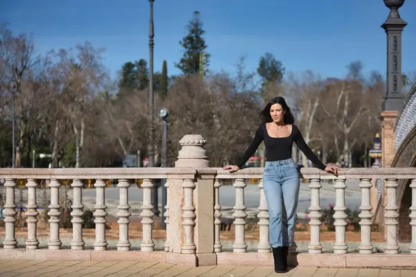Genç, güzel, esmer bir kadın, siyah uzun kollu tişört ve kot pantolon giyiyor, saçları rüzgarda uçuşuyor, Seville 'de İspanya meydanında bir korkuluğa yaslanmış poz veriyor. Güzellik kavramı, kadınlık.