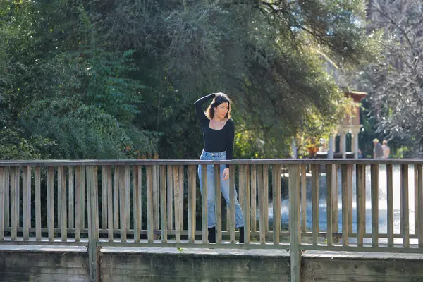 Siyah tişört ve kot pantolon giymiş, genç, güzel, esmer bir kadın. Tahta bir köprüde fotoğraf çekilirken poz veriyor. Güzelliği, modayı, eğilimi, kadınlığı kavra.