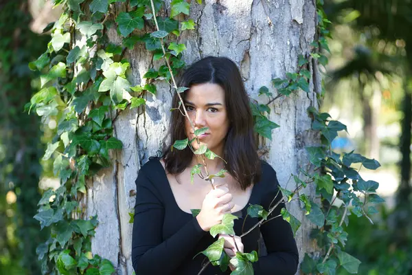 Siyah tişört ve kot pantolon giymiş, bir ağacın gövdesine yaslanmış ve doğayla bağlantılı, sarmaşığa dolanmış genç, güzel, koyu renk saçlı bir kadın. Güzellik kavramı, kadınlık, doğa, bitkiler, ağaçlar.