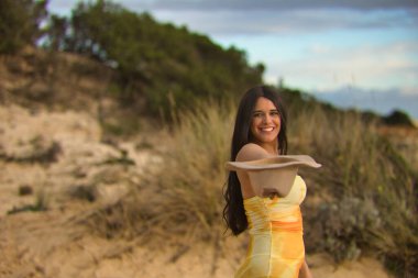 Genç, güzel, esmer, sarı elbiseli, elinde şapkasıyla kameraya bakıp gülümseyen ve eğlenen bir kadın. Güzelliği, modayı, eğlenceyi, mutluluğu, hayatı kavra.