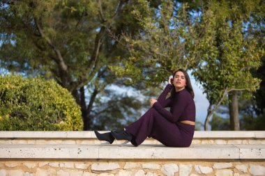 Genç, güzel, esmer, İspanyol bir kadın, mor elbiseli ve topuklu ayakkabılı, duvarda oturuyor, kameraya şefkatli ve hayalperest bakıyor. Güzelliği, modayı, modayı kavra.