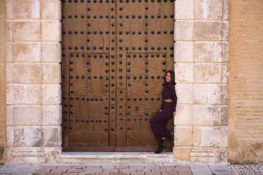 Genç, güzel, esmer, İspanyol bir kadın, mor bir elbise ve topuklu ayakkabı giyiyor, kameraya bakıyor, büyük, eski bir ahşap kapının çerçevesine yaslanıyor. Güzelliği, modayı, modayı kavra.