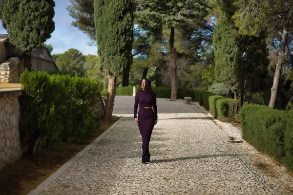 Genç, güzel, esmer, İspanyol bir kadın, mor bir elbise ve yüksek topuklu ayakkabı giyiyor, selvi ağaçlarıyla çevrili taş bir yolda tek başına yürüyor. Güzelliği, modayı, modayı kavra.