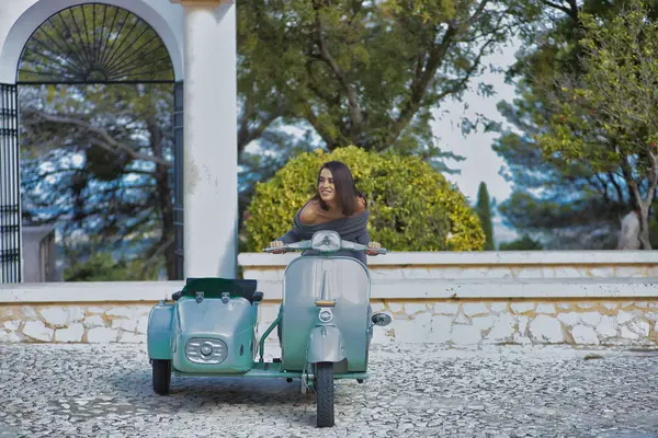 Genç, güzel, esmer, gri elbiseli İspanyol kadın sepetli klasik bir motosiklete biniyor, çok mutlu ve memnun. Motor konsepti, motosikletler, vintage, moda, güzellik.