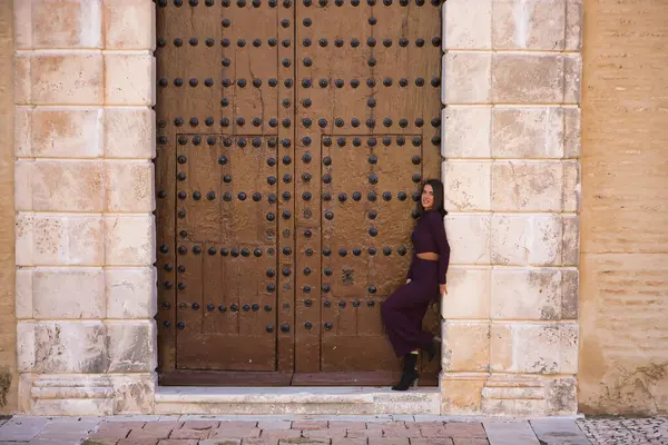 Genç, güzel, esmer, İspanyol bir kadın, mor bir elbise ve topuklu ayakkabı giyiyor, kameraya bakıyor, büyük, eski bir ahşap kapının çerçevesine yaslanıyor. Güzelliği, modayı, modayı kavra.