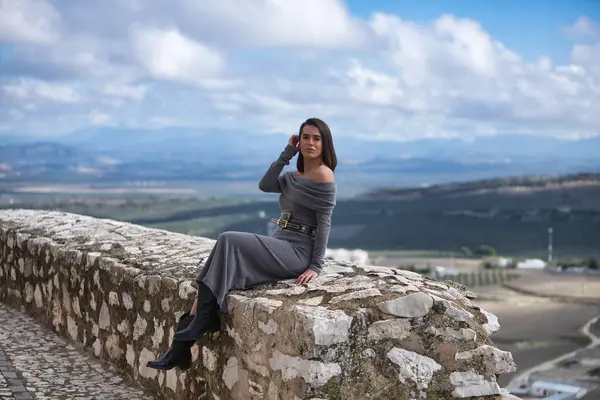 Genç, güzel, esmer, İspanyol kadın, gri bir elbise ve çizme giyiyor, sonsuzluğa bakıyor, taş bir duvarda oturuyor, arkasında bulutlardan ve kırsal alanlardan oluşan güzel bir manzara var..