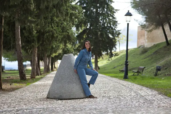Genç, güzel, esmer, İspanyol bir kadın, kot elbise giyiyor, gülümsüyor ve mutlu görünüyor, taş bir yolda eski bir değirmen taşına yaslanıyor. Kavram moda, güzellik, moda, kadınlık.