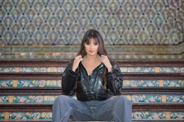 Genç, güzel, esmer kadın siyah timsah derisi ceket ve kot pantolon giymiş, kameraya bakıyor, modern ve moda, İspanya 'nın Seville meydanında bir merdivende oturuyor..