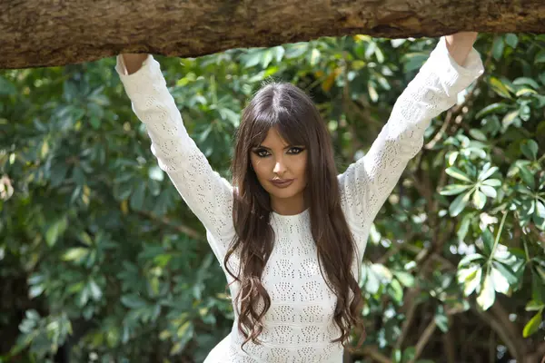 Genç, güzel, esmer bir kadın, beyaz örgü bir elbise giyiyor, kameraya bakıyor, elleri bir ağaç gövdesinde dinleniyor, sırtında güneş ışınları alıyor..