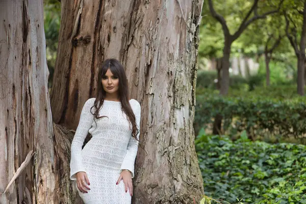 Beyaz örgü elbiseli, genç, güzel, esmer kadın, kameraya bakıyor, doğayla bağlantılı, büyük bir ağacın gövdesine yaslanıyor..