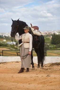 Genç bayan binici, siyah İspanyol safkan atının yanında poz veriyor, dizginleri elinde tutuyor. Konsept at biniciliği, atlı kadın, at biniciliği, binicilik. Dikey konum.