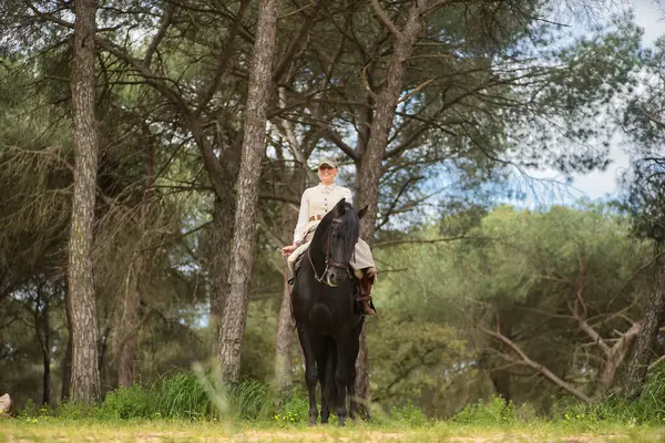 Siyah İspanyol safkan atına binen genç atlı kadın, tarlanın ortasında poz veriyor. Yanında bir eyer ve elinde bir kırbaç var. Konsept at biniciliği, at biniciliği, binicilik, binicilik.