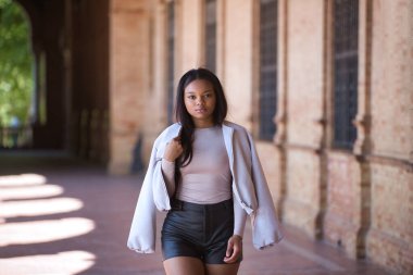 Genç, güzel, Afrikalı bir kadın, ceketli ve deri şortlu, İspanya meydanında poz veriyor ve modellik yapıyor. Moda konsepti, güzellik, moda.