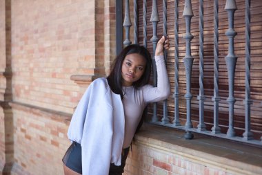 Genç, güzel, deri ceketli ve şortlu Afrikalı bir kadın. Sevilla 'da İspanya meydanındaki bir pencerenin ızgarasına tatlı ve yumuşak bir bakış atıyor. Moda konsepti, güzellik, moda.