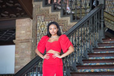 Genç, güzel, Afrikalı bir kadın, kırmızı bir elbise giyiyor, Sevilla 'da İspanya meydanının merdivenlerinde elleri belinde bir model gibi poz veriyor. Moda konsepti, güzellik, moda.