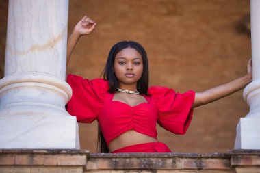 Genç, güzel, kırmızı elbiseli Afrikalı bir kadın Sevilla 'da İspanya meydanında iki mermer kolonun arasına yaslanmış kameraya bakıyor. Moda konsepti, güzellik, moda.