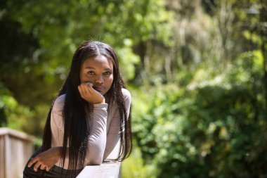 Genç, güzel, Afrikalı bir kadının portresi, tatlı ve yumuşak bakışlı, çenesinde bir el ve açık hava parkındaki ahşap bir korkulukta dinleniyor. Güzellik kavramı, görünüş.