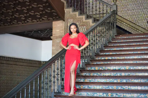 Genç, güzel, Afrikalı bir kadın, kırmızı bir elbise giyiyor, Sevilla 'da İspanya meydanının merdivenlerinde elleri belinde bir model gibi poz veriyor. Moda konsepti, güzellik, moda.