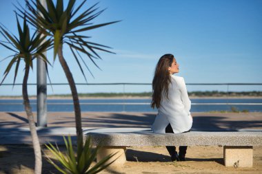 Genç, güzel, kahverengi saçlı, beyaz ceketli bir kadın, sırtı dönük, bir bankta oturuyor, yalnız ve sessiz, şafak vakti güneşin ışınlarını alıyor..
