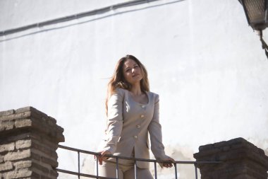 Açık gri ceketli, genç, güzel, kahverengi saçlı bir kadın kameraya bakıyor, korkuluklara yaslanıyor, aşağıdan görünüyor..