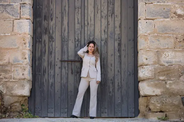 Genç, güzel, kahverengi saçlı bir kadın, açık gri bir ceket giyiyor, kameraya bakıyor, saçlarına dokunurken poz veriyor, kocaman, ahşap bir kapının ortasında..