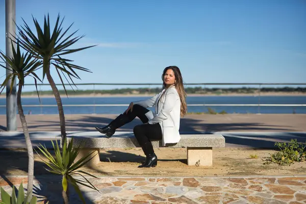 Genç, güzel, kahverengi saçlı, beyaz ceketli bir kadın, kameraya bakıyor, bir bankta yan oturuyor, yalnız ve sessiz, şafak vakti güneş ışınlarını alıyor..