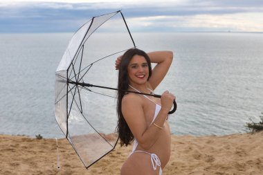 Genç, güzel, esmer, beyaz bikinili kadın, şeffaf bir şemsiyeyle poz veriyor, arka planda deniz var. Güzellik, doğa, plaj, deniz, banyo modası.