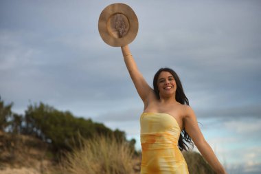 Sarı elbiseli, genç, güzel, koyu renk saçlı bir kadın, bir kolu havada, elinde şapkasıyla, gülümseyen ve eğlenen kameraya bakıyor. Güzelliği, modayı, eğlenceyi, mutluluğu, hayatı kavra.