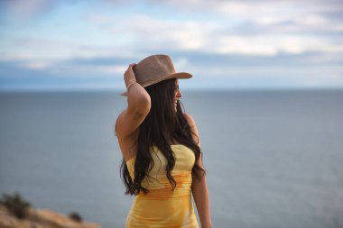 Sarı elbiseli, siyah saçlı, genç bir kadın gün batımında denize bakıyor. Güzellik, moda, plaj, deniz, gün batımı.