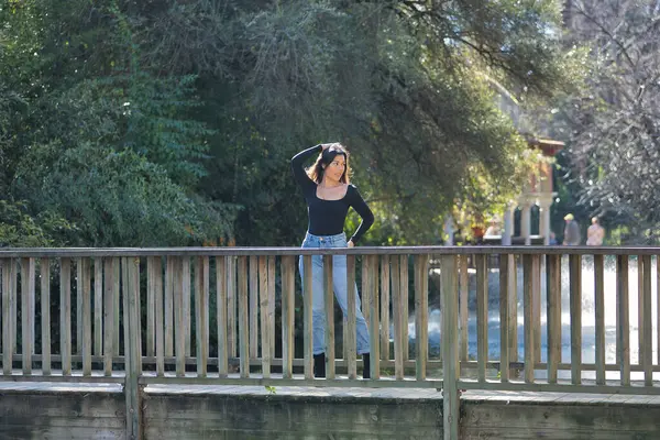 Siyah tişört ve kot pantolon giymiş, genç, güzel, esmer bir kadın. Tahta bir köprüde fotoğraf çekilirken poz veriyor. Güzelliği, modayı, eğilimi, kadınlığı kavra.