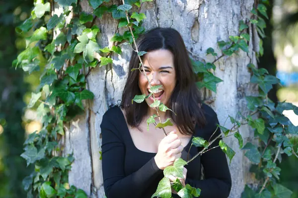 Genç, güzel, esmer bir kadın, siyah tişört ve kot pantolon, bir ağacın gövdesine yaslanmış, sarmaşıkla komik oynuyor, doğayla bağlantılı. Güzellik kavramı, kadınlık, doğa, bitkiler, ağaçlar.