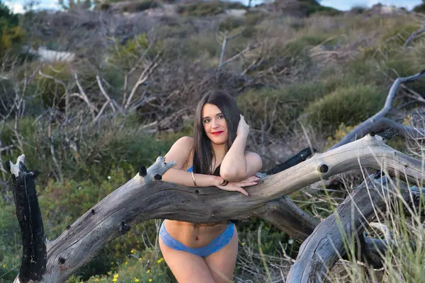 Genç bir kadın, güzel, esmer, mavi iç çamaşırlı, kameraya flört ederek bakıyor ve bir ağaç gövdesine yaslanmış gülümsüyor. Güzelliği kavra, doğayı, modayı, flörtü.