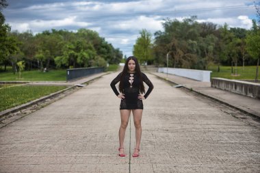 Genç bir kadın, esmer, güzel, soluk tenli, siyah bir elbise ve yüksek topuklu giyen, açık hava parkının ortasında elleri belinde poz veren..